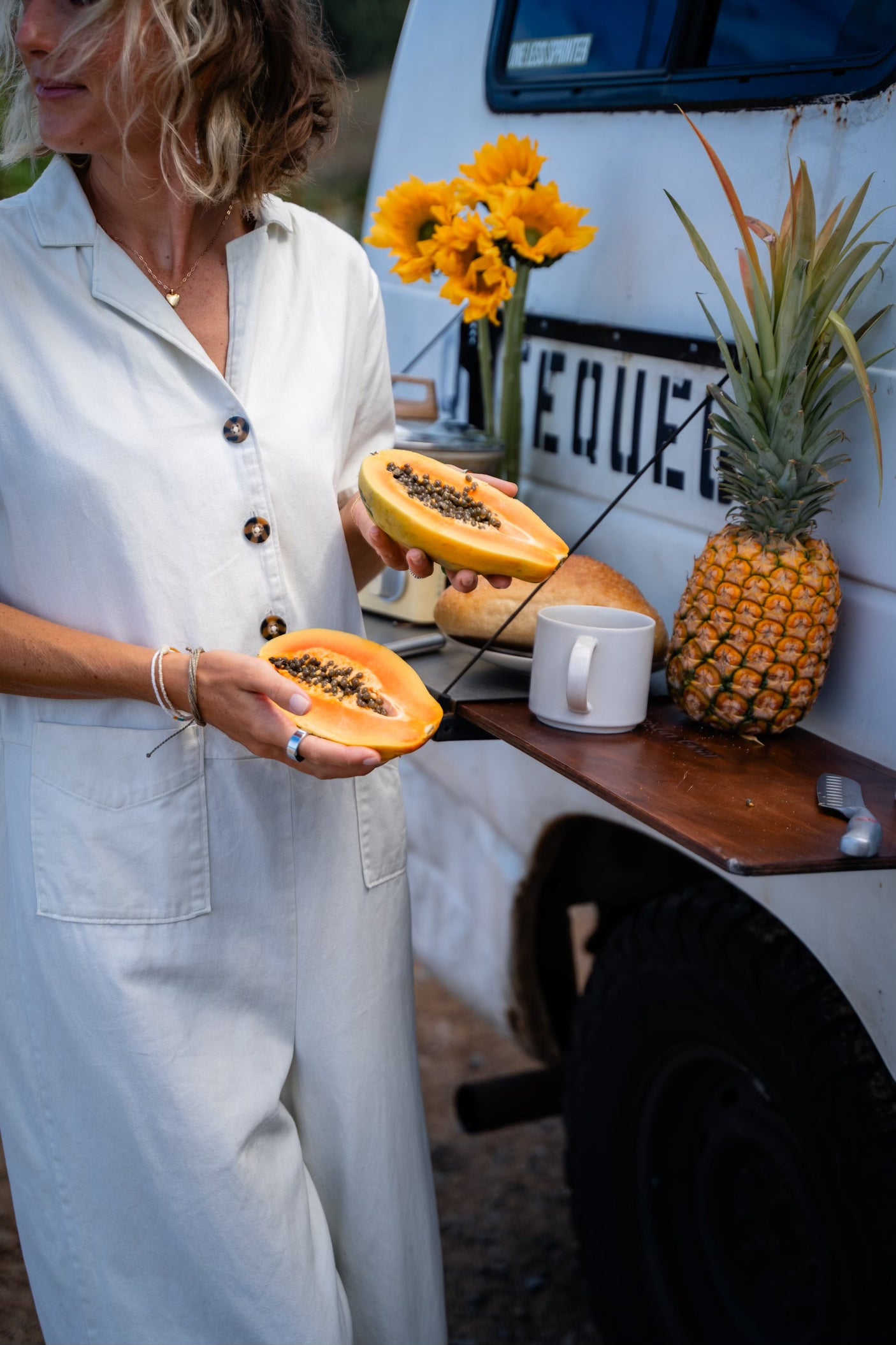 A woman holds fresh papaya near an RV rental, set up for a healthy breakfast with pineapple, bread, and sunflowers. Perfect for van life.