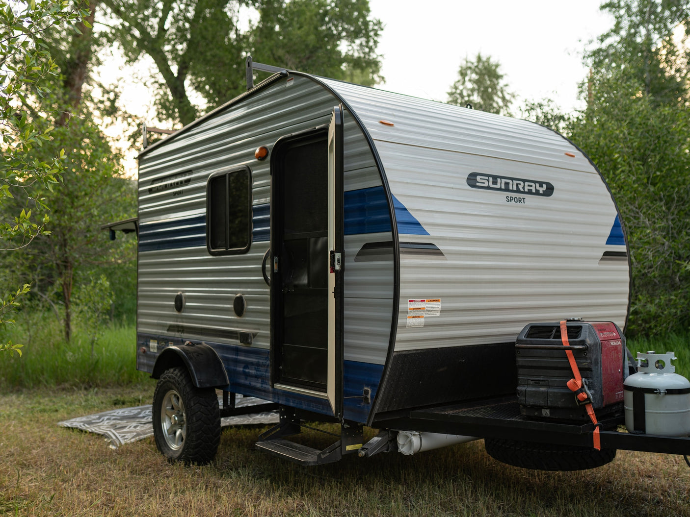 A Sunray Sport travel trailer rv rental in a scenic campsite, featuring an open door, generator, and propane tank.