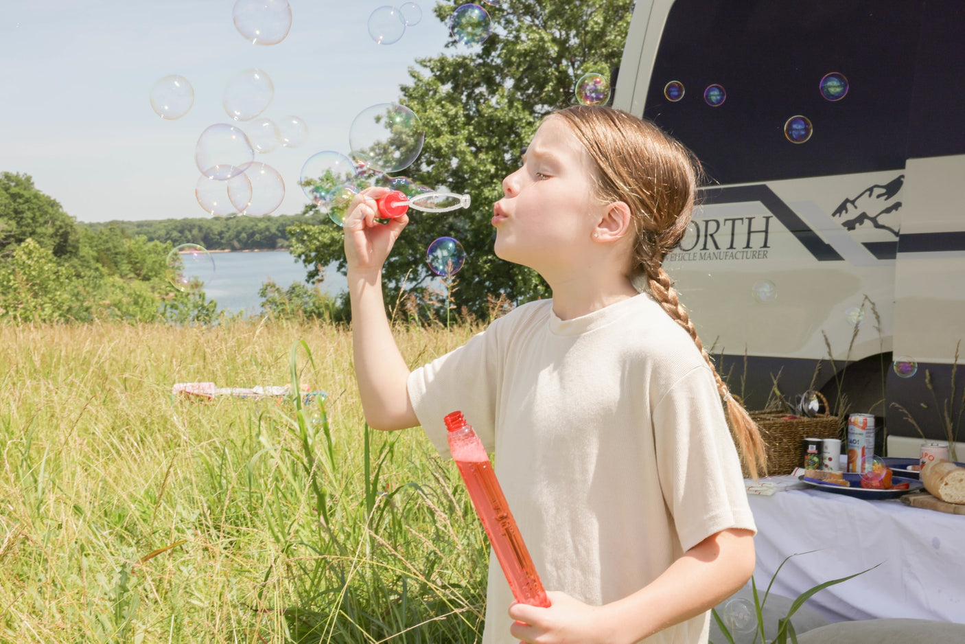 A young girl blows colorful bubbles outdoors by an RV rental, with a lake and trees in the background.
