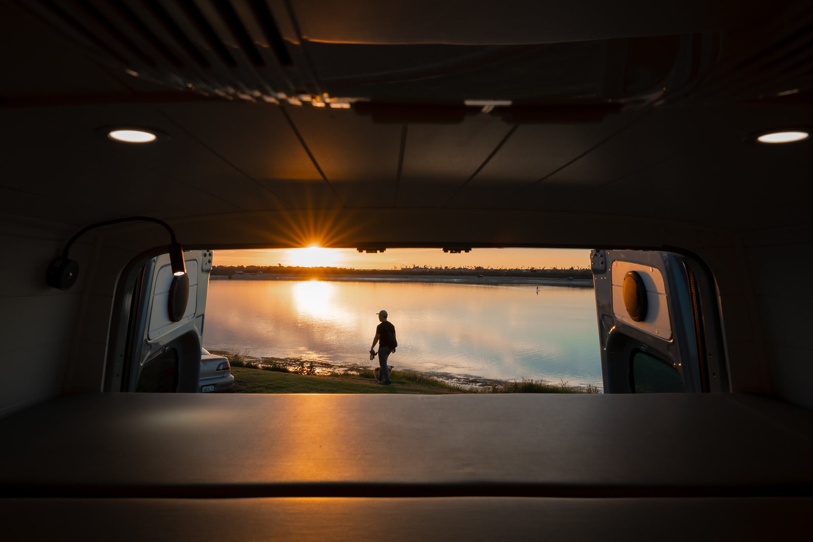 Panoramic sunset from an RV rental. A man & dog stand by shimmering water, seen from inside the cozy van.