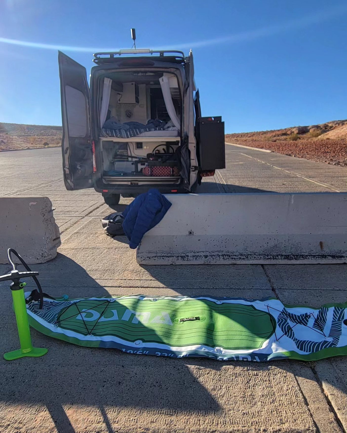 An RV rental camper van with open doors on a desert road. A paddleboard and pump lie ready for adventure in the foreground.