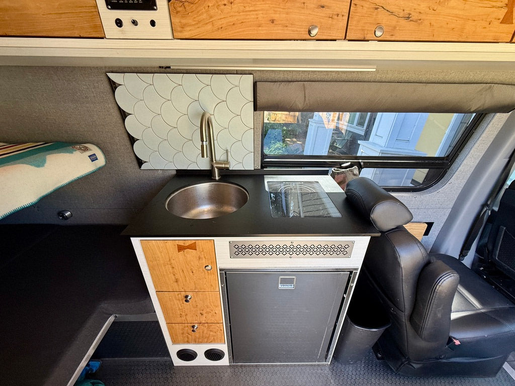 Modern RV rental interior: a sleek kitchenette with sink, cooktop, fridge, wood cabinets, and cozy bed area for travel.