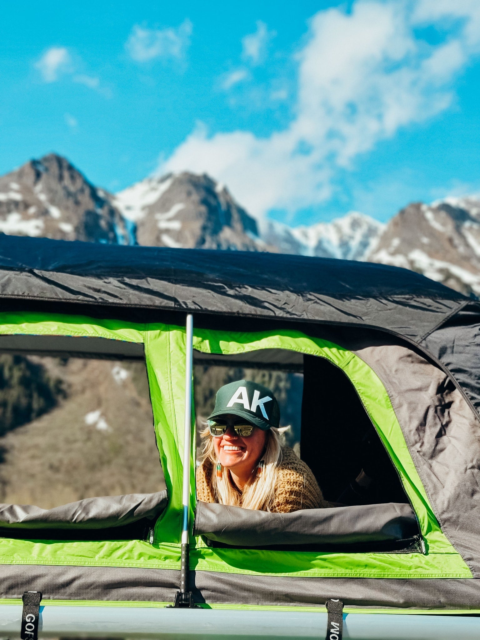Smiling woman in an RV rental rooftop tent enjoys majestic mountain views on a sunny day.
