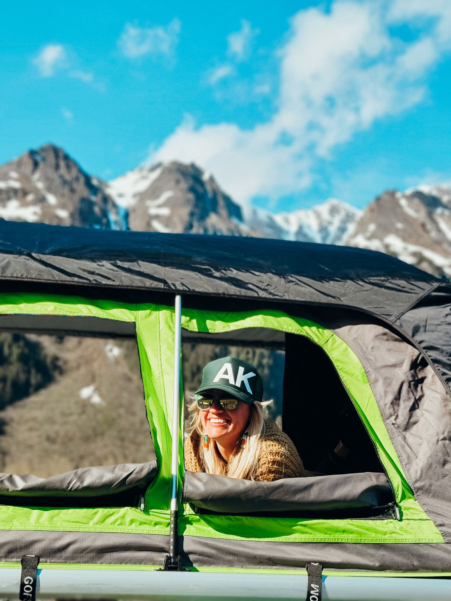 Smiling woman in an RV rental rooftop tent enjoys majestic mountain views on a sunny day.