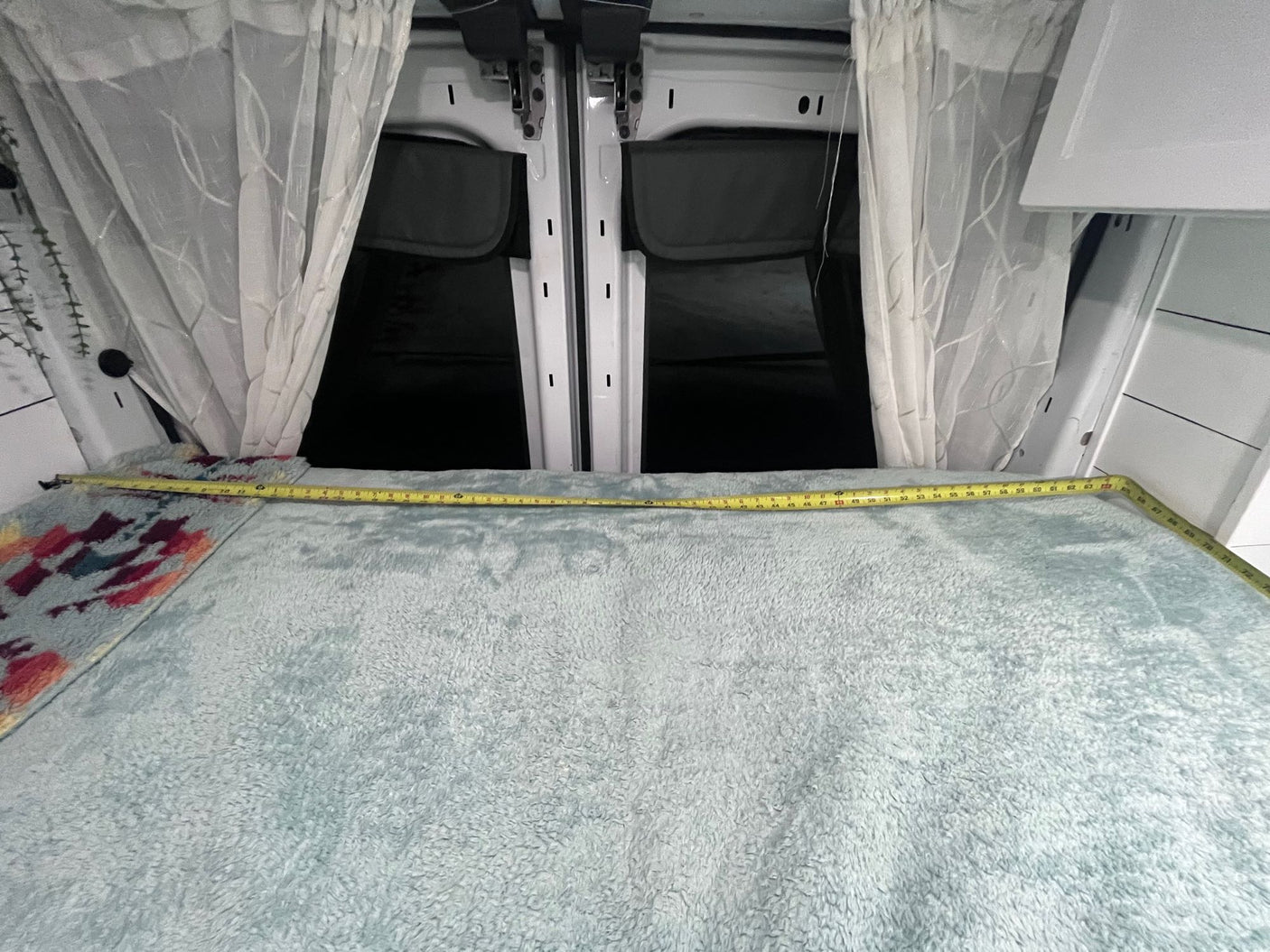 Cozy RV rental sleeping area: Bed with measuring tape, sheer curtains frame rear van doors.