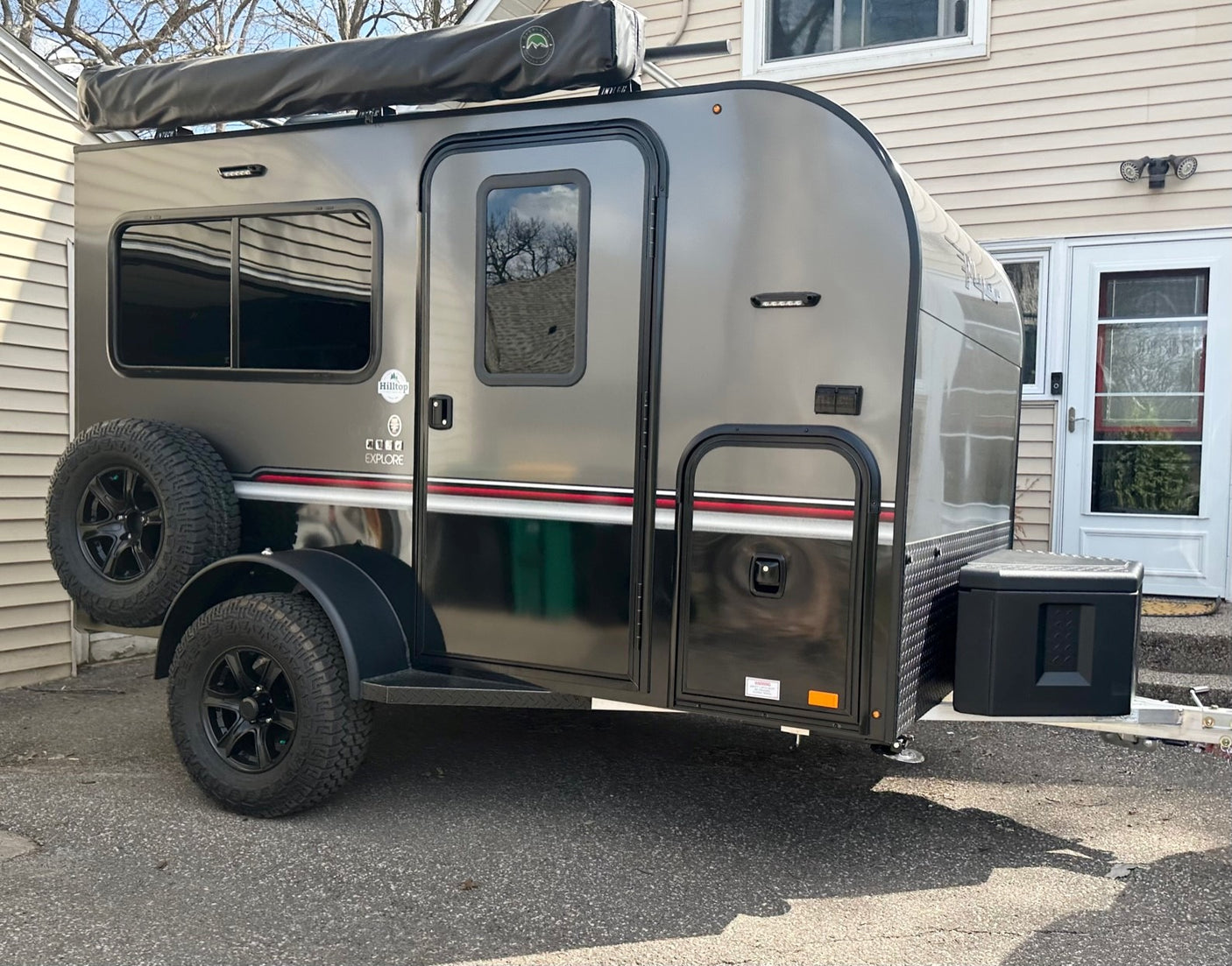 Compact, dark gray travel trailer/teardrop RV for rental. Features include an awning, rugged tires, and a spare. Ready for adventure.