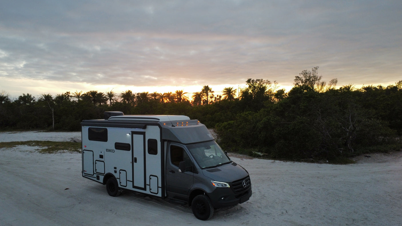 RV rental: A grey Mercedes-Benz camper van parked on sandy terrain at sunrise/sunset, with a warm sky glow.