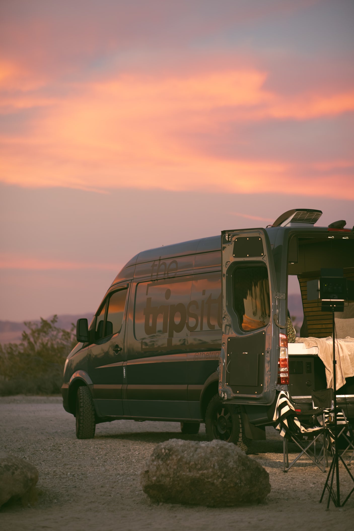 This 'the tripsit' RV rental camper van, parked at sunset, has its rear doors open, revealing a cozy interior in a desert setting.