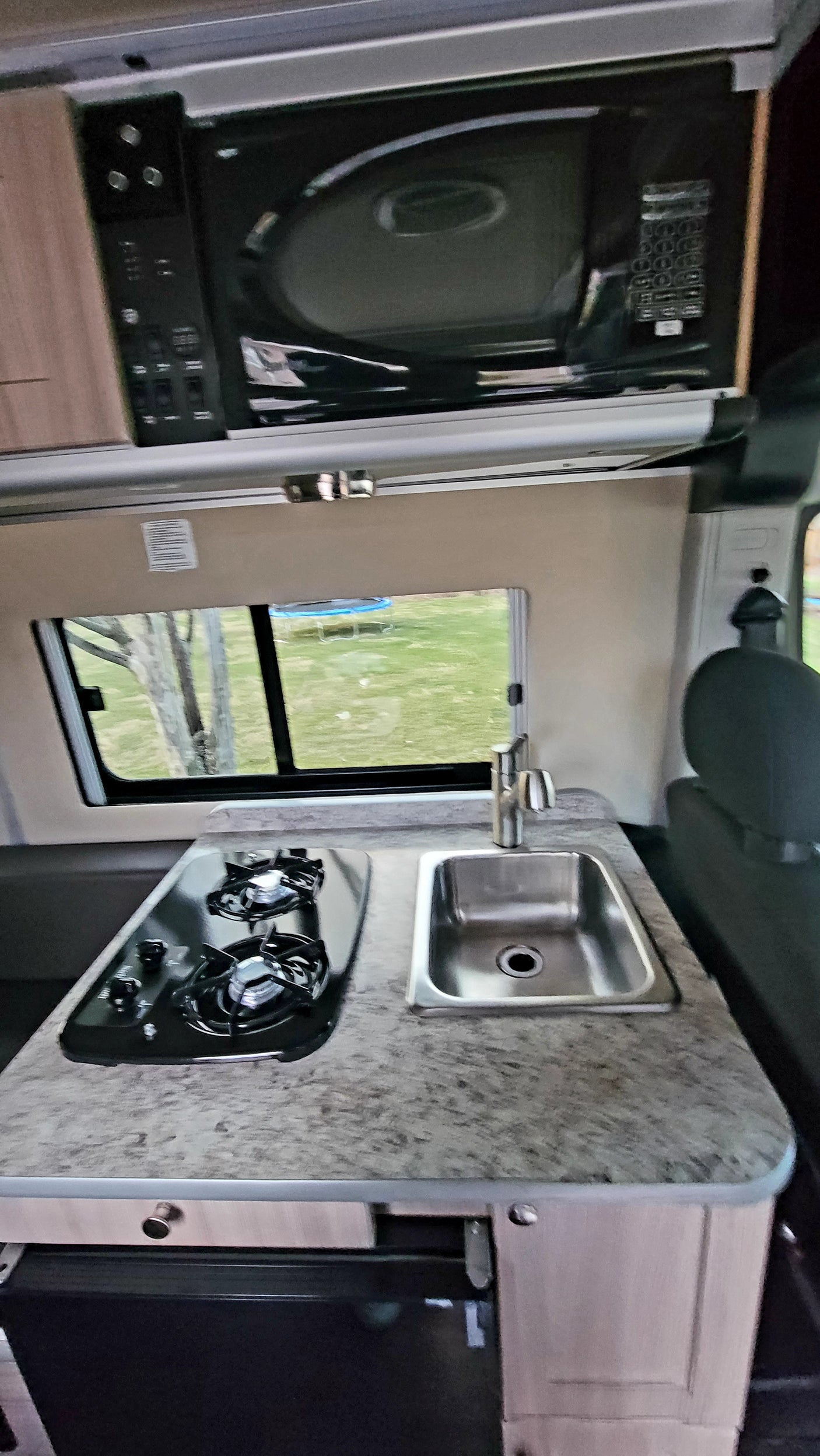 Compact RV rental kitchenette: microwave, two-burner stove, and sink. Window view of outdoors.