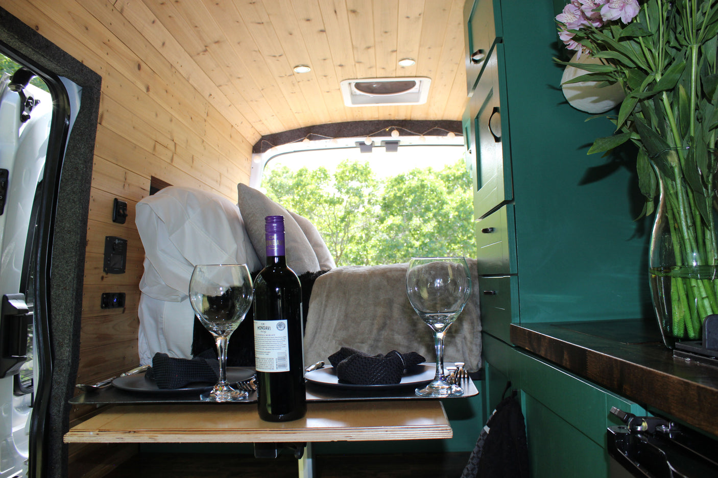 Romantic RV rental interior showcases wooden walls, teal cabinets, and a table set for two with wine, flowers, and a scenic view.