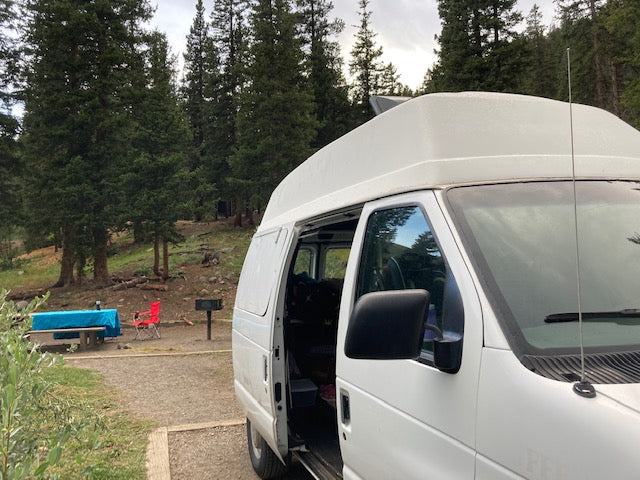 RV rental camper van parked at a scenic campsite with picnic table and grill among tall trees.