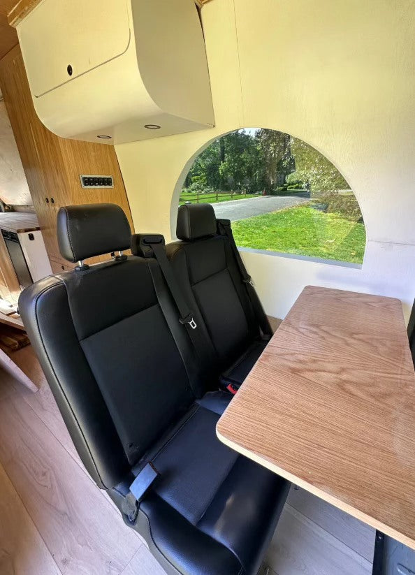 RV rental interior: cozy camper van with black seats, wooden table, overhead storage, and an arch window view.