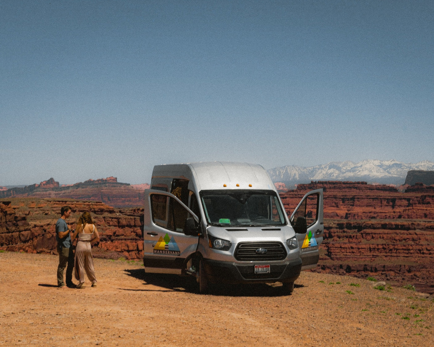 A couple stands by their silver RV rental van in a vast desert landscape, with red rock formations and snowy mountains.
