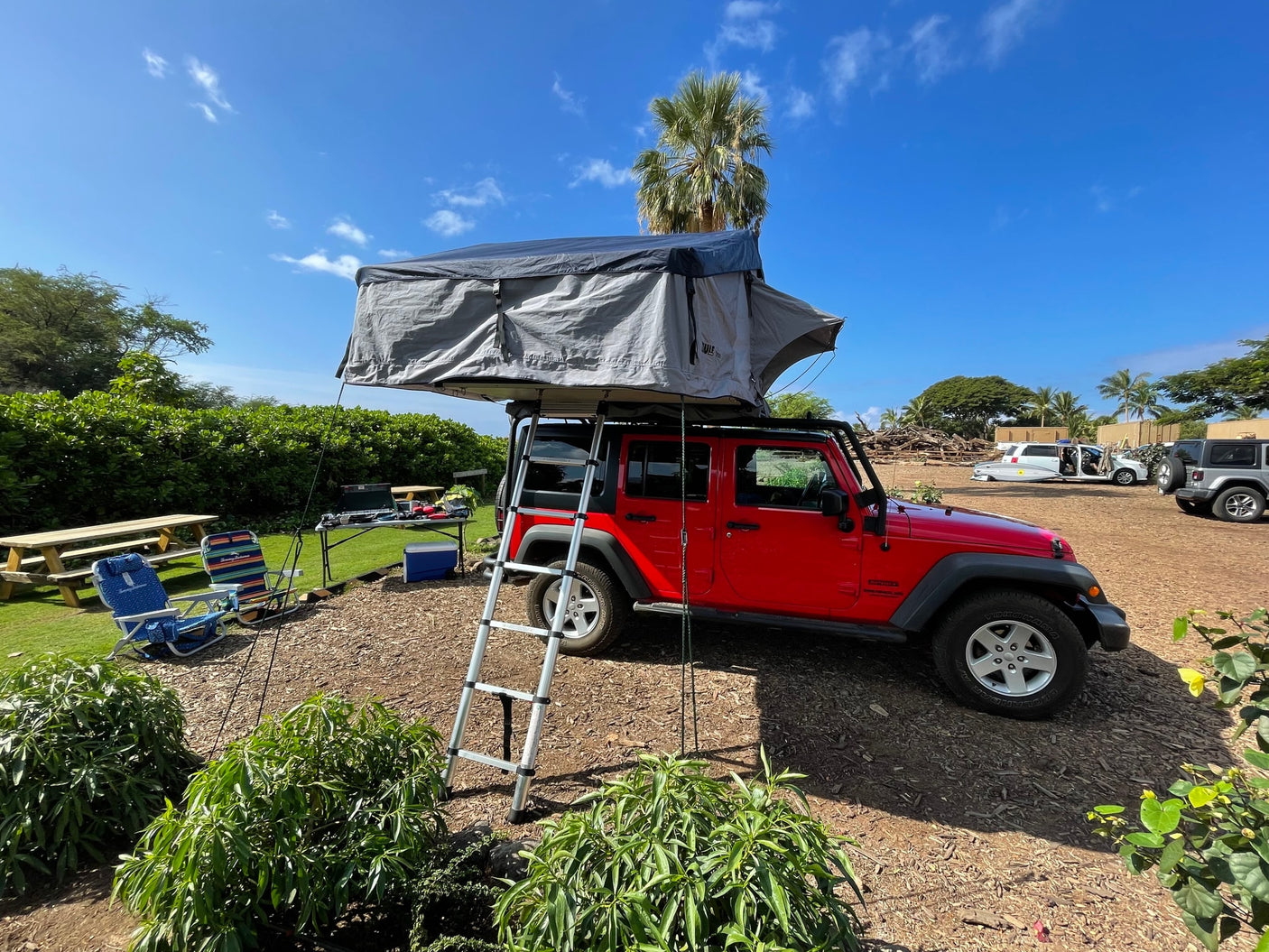 Red Jeep Wrangler with grey rooftop tent, ladder extended, set up at a sunny outdoor camp. Perfect for an RV rental trip.