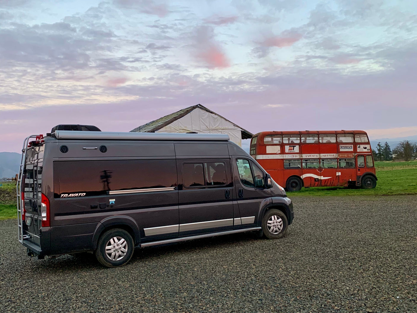 Dark gray RV rental parked on gravel near an old red double-decker bus and a white structure under a vibrant sky.