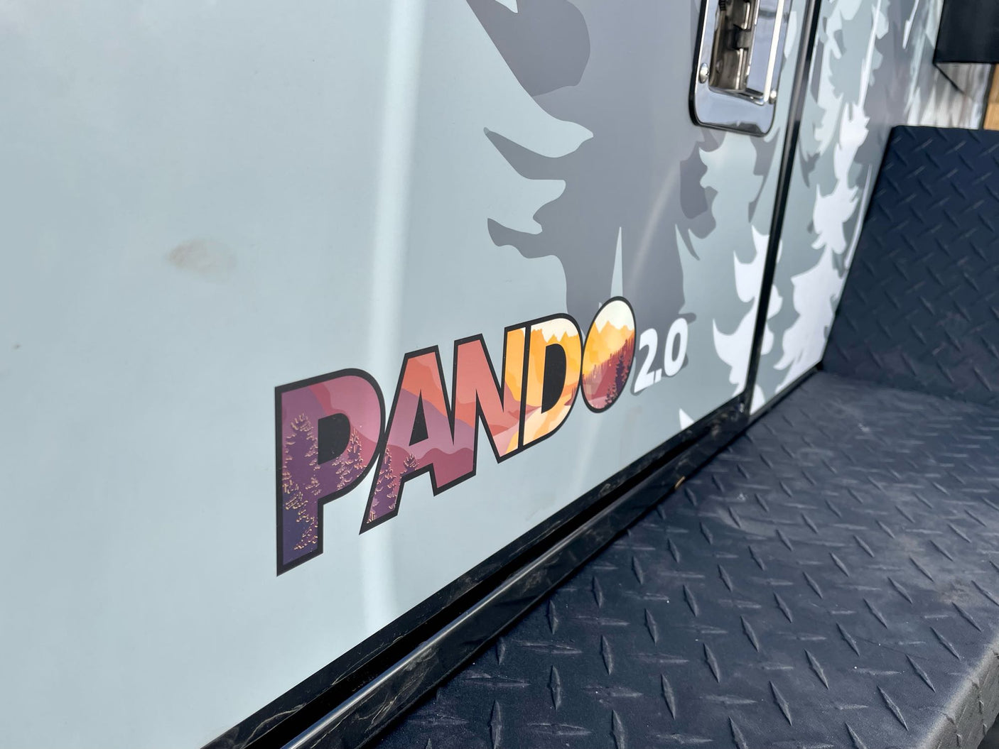 Side of PANDO 2.0 RV rental: mountain-filled logo, forest camouflage, and black diamond plate. Ready for adventure.