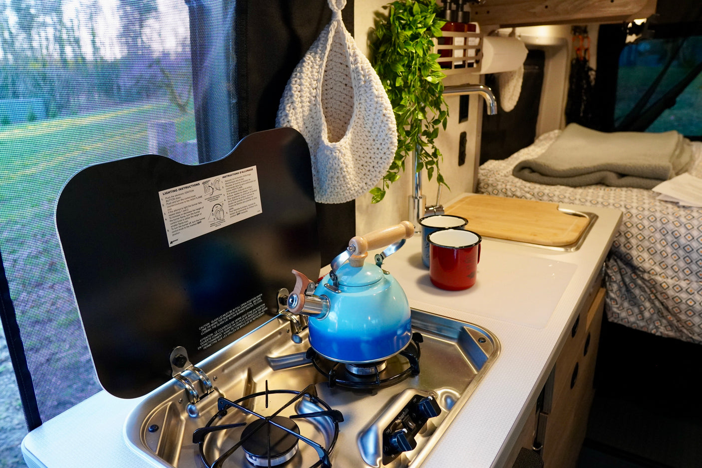 Compact RV rental kitchen with stove, kettle, sink, and comfy bed beyond. Perfect for mobile living.
