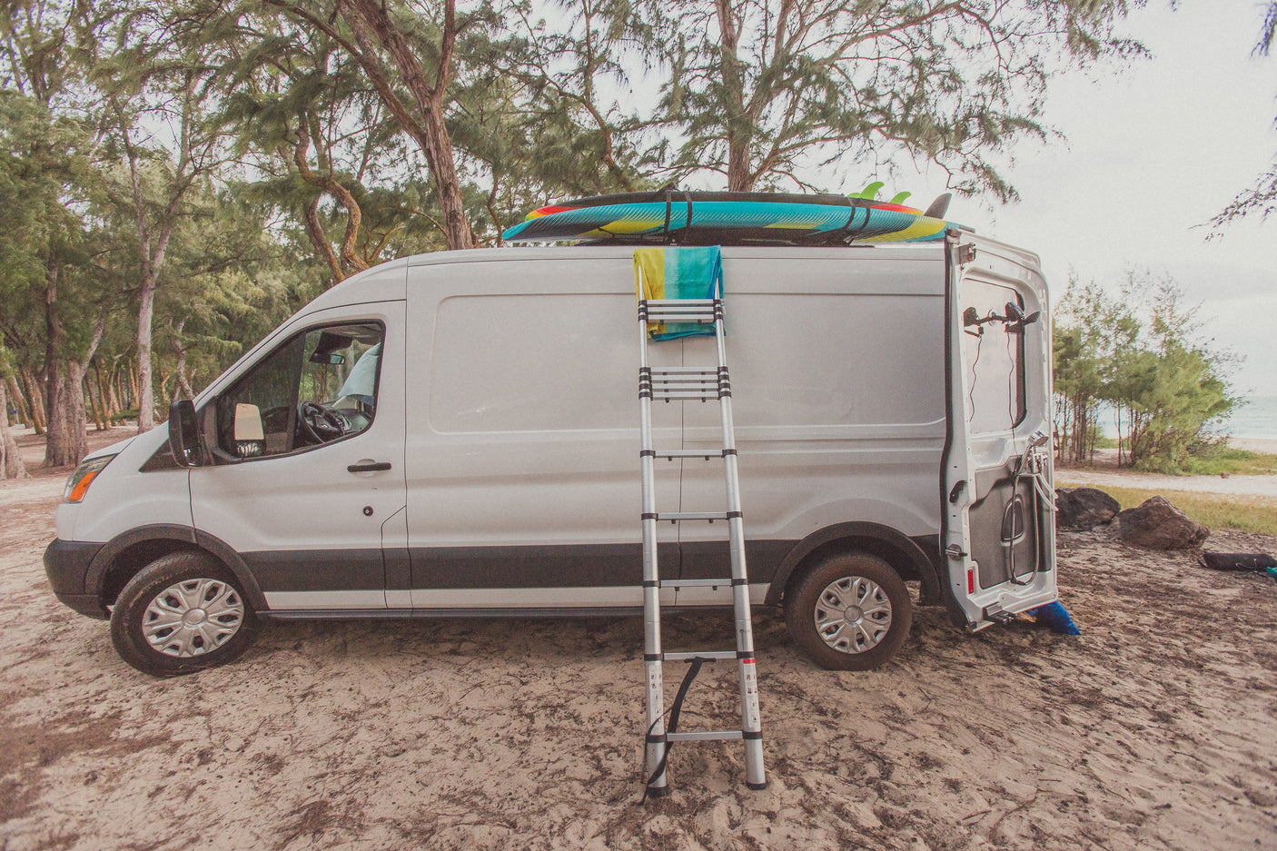 White RV rental van on sandy ground with surfboards, ladder, and a beach shower setup. Ready for coastal adventures!