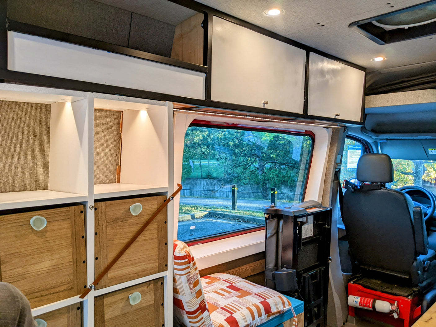 Bright RV rental interior: custom wooden storage, white overhead cabinets, a large window view, and a fire extinguisher.