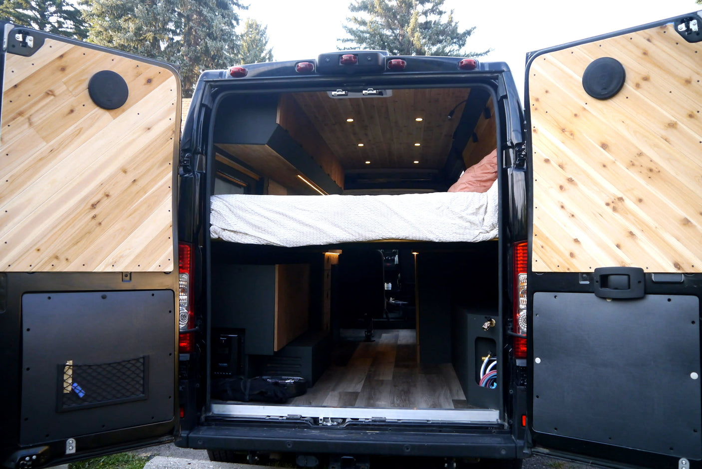 Open rear doors of an RV rental camper van, revealing its custom wood interior with a bed and storage space.