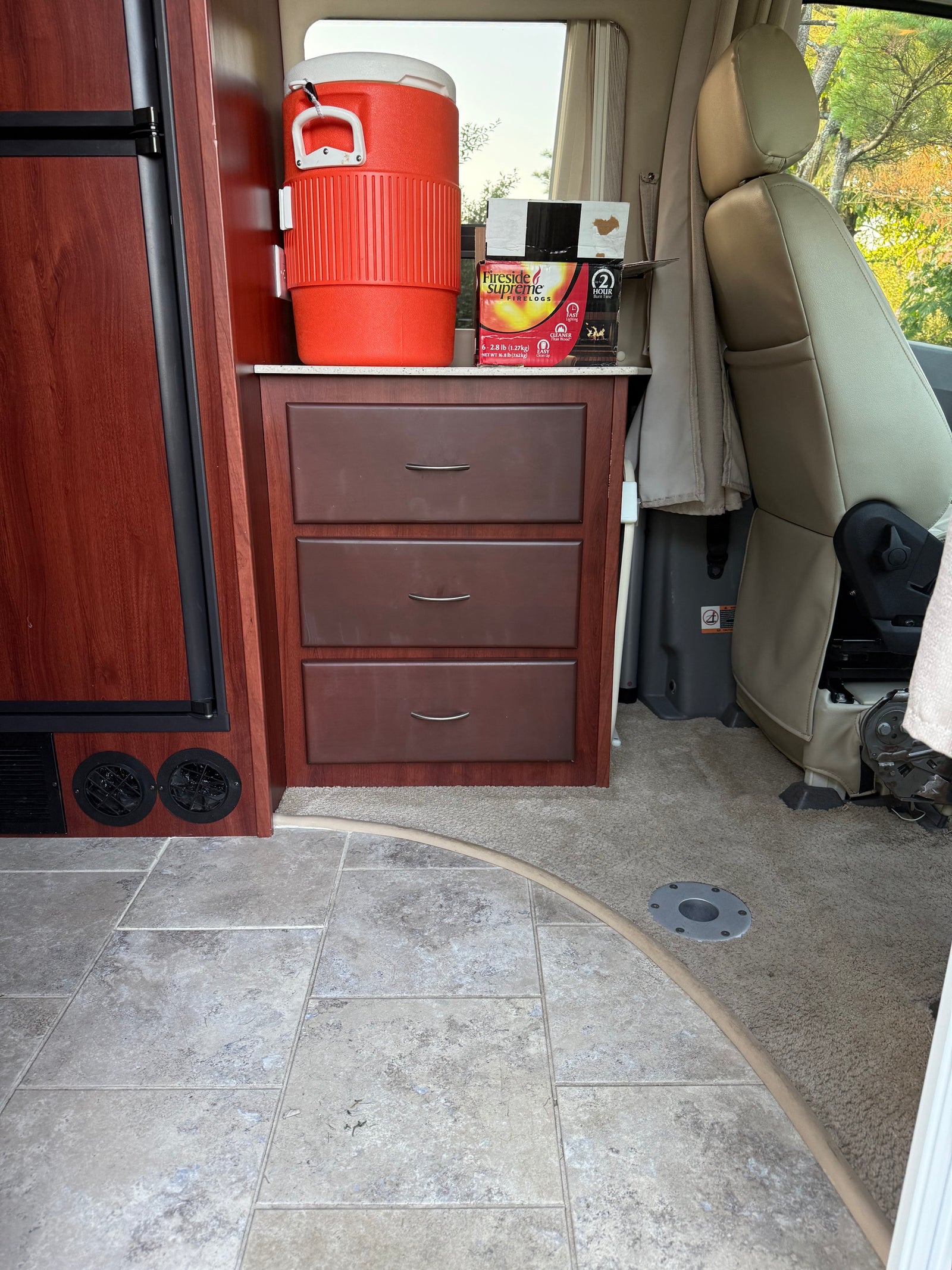 RV rental interior view of functional kitchen cabinetry, a large cooler, firelogs, and front passenger seating.