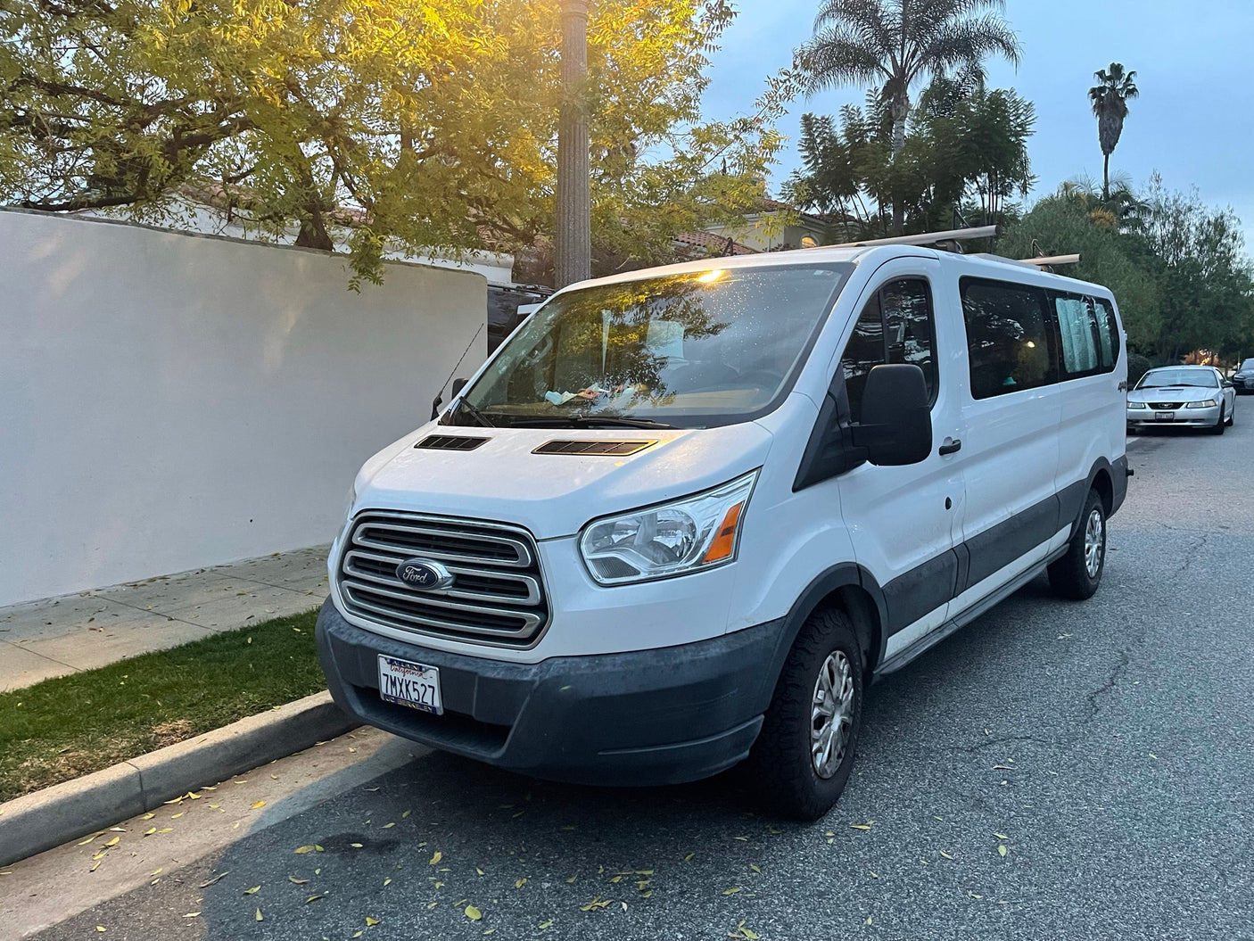A white Ford Transit passenger van, often used as an rv rental, parked on a tree-lined California street. Plate 7MXK527.
