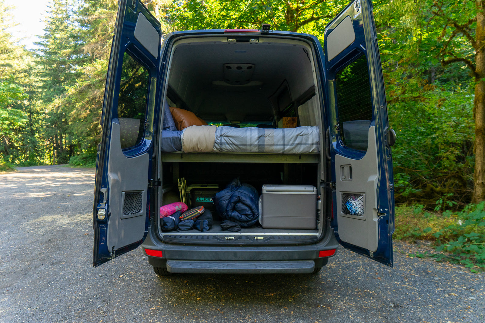 Open rear doors of a blue RV rental reveal a comfortable bed above and ample gear storage below for your next van life adventure.
