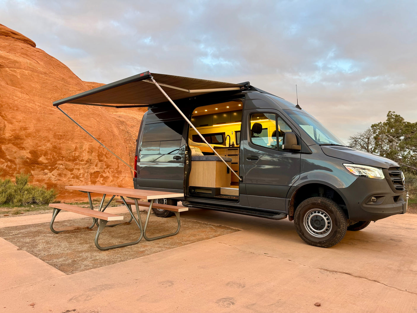 Modern rv rental camper van with extended awning & inviting interior, parked with a picnic table by red rock formations.