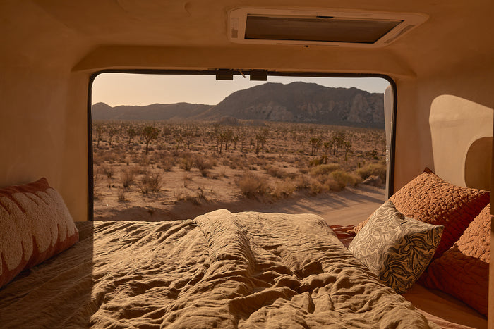 Inside an rv rental: a comfy bed with warm blankets, framing a window view of a desert, Joshua trees, & mountains.