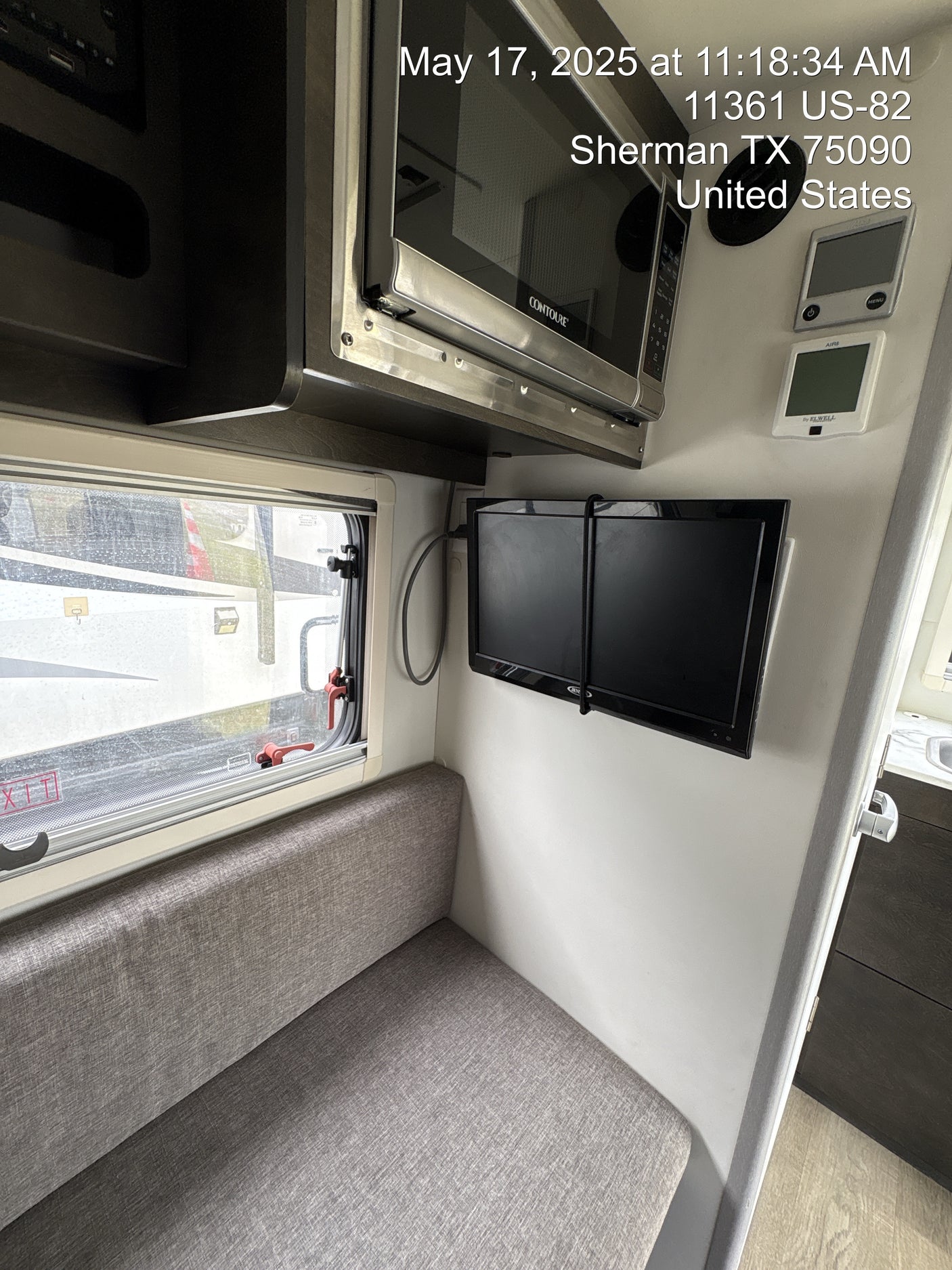 View of a modern rv rental interior: gray upholstered seating, wall-mounted TV, microwave, and window showing exterior.