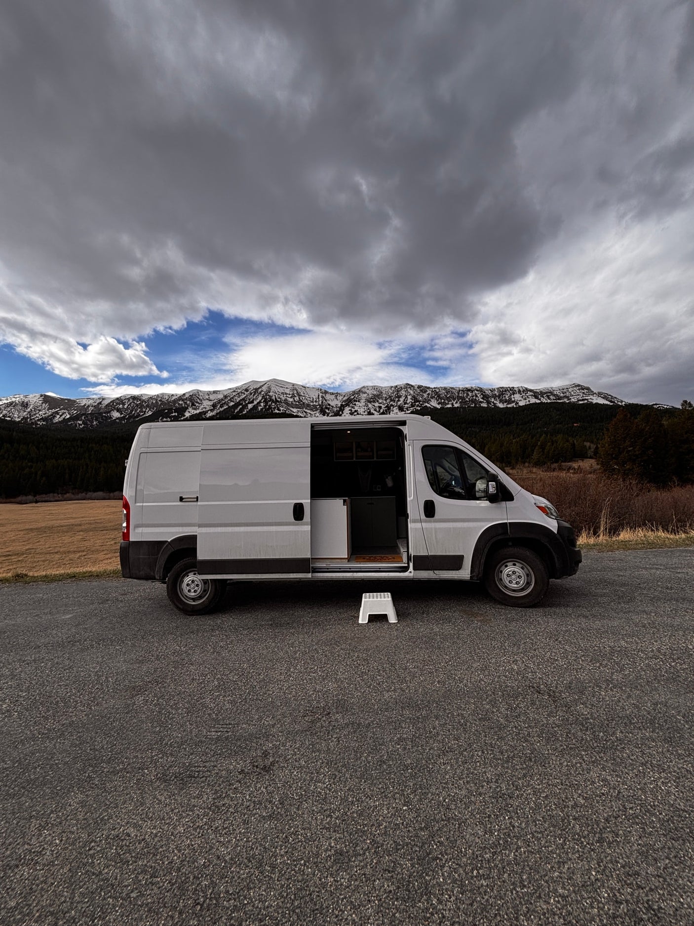 White RV rental camper van with open side door, step stool, parked by snowy mountains under a cloudy sky.