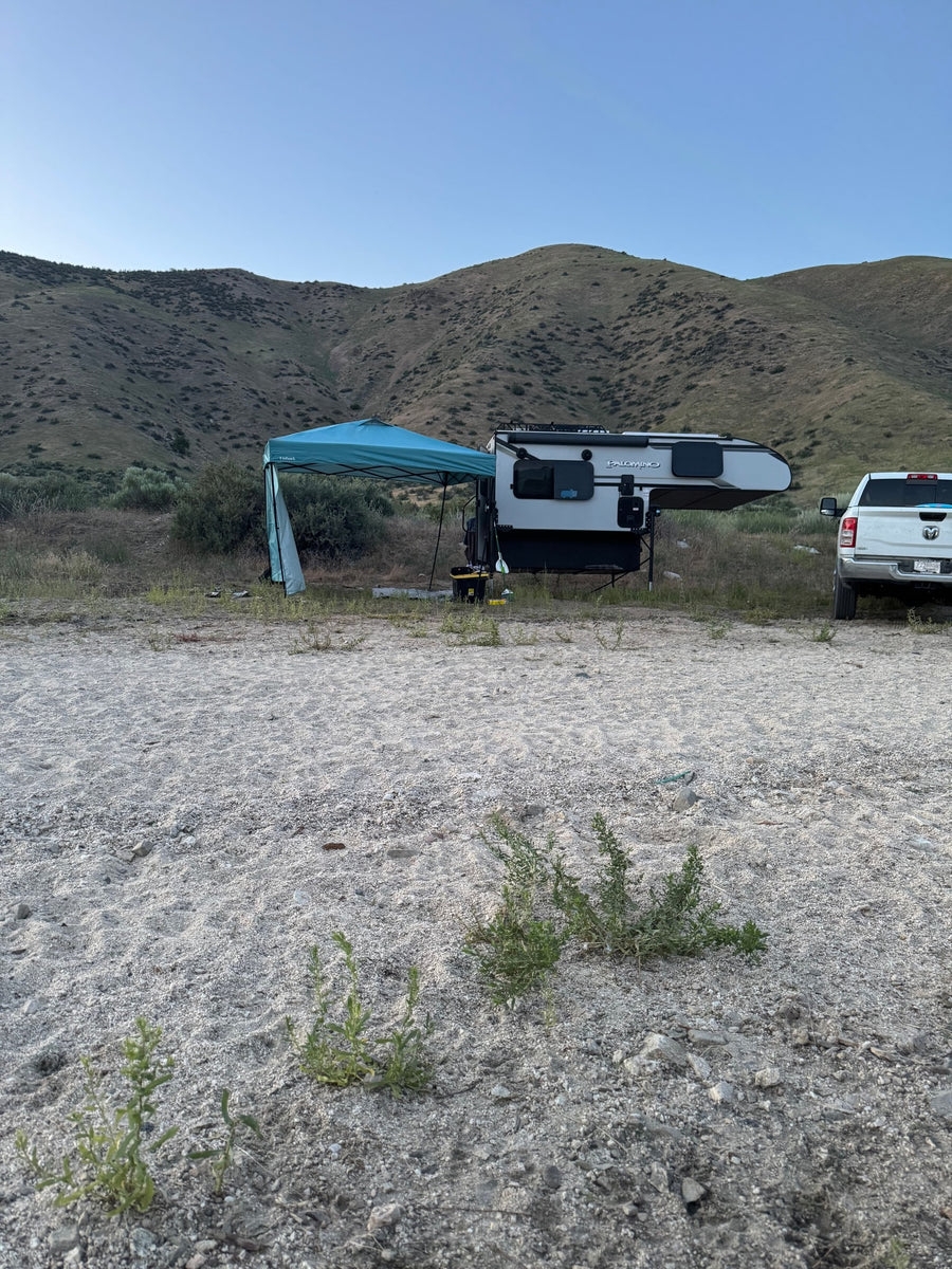 A grey truck with an RV rental truck camper & a blue canopy set up at a gravel campsite, backed by vegetated hills.
