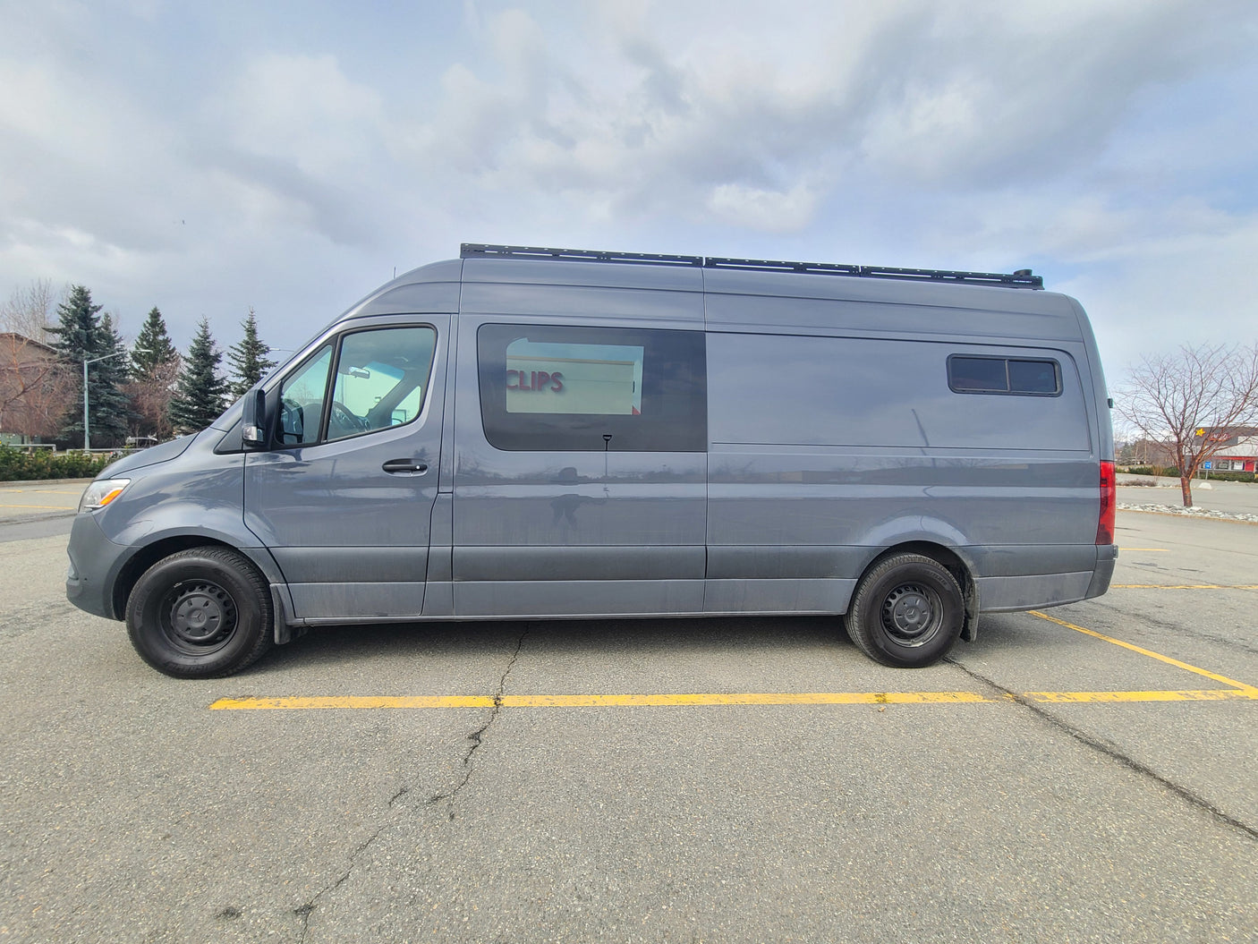 This gray Sprinter camper van, featuring a roof rack, is ideal for an RV rental, parked in a lot under cloudy skies.