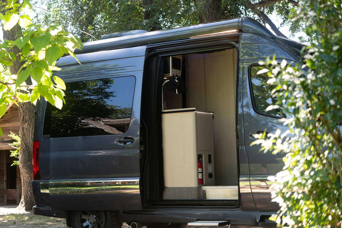 An open door reveals the modern interior of this dark gray RV rental, featuring light wood cabinetry, a sink, and a fire extinguisher. — photo 8