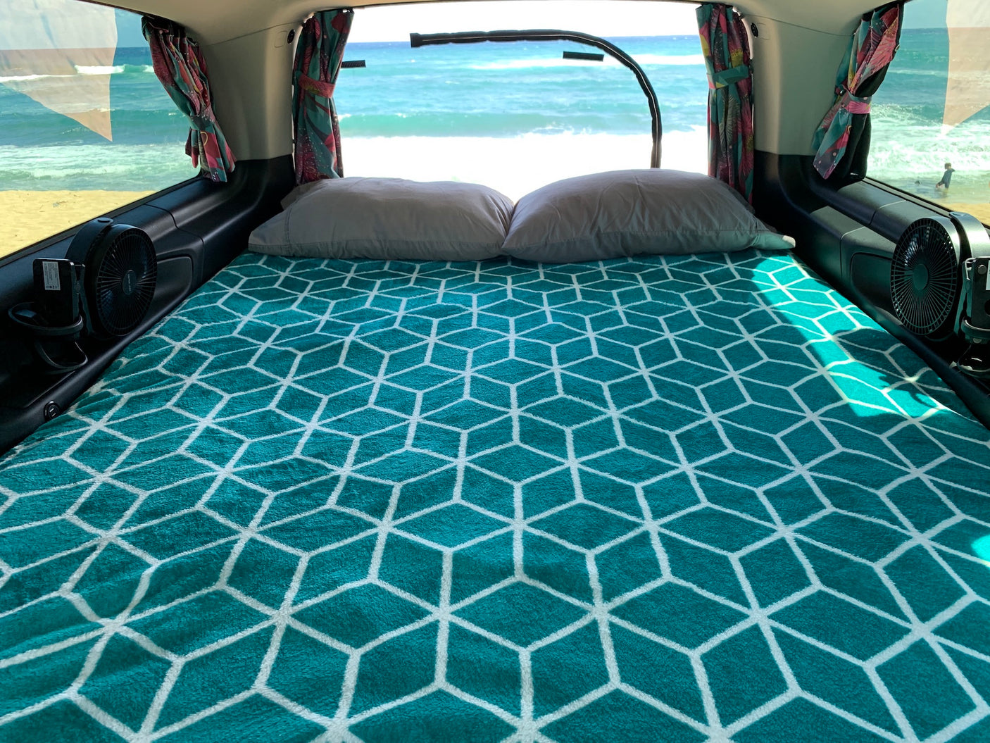 Cozy **rv rental** camper bed with teal geometric blanket, pillows, and fans, enjoys a direct ocean view from inside.
