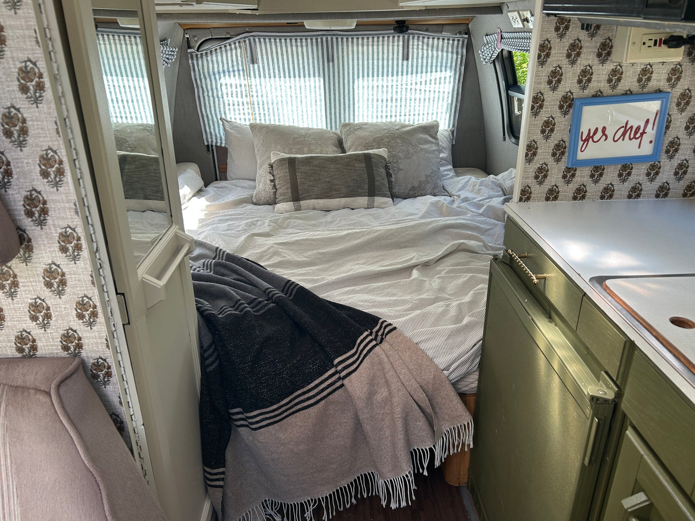 Cozy RV rental interior showcasing a comfortable bed with striped bedding, curtains, and a compact kitchen with a "yes chef!" sign.