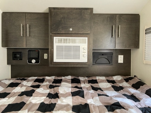 This RV rental's bedroom shows a dark wood headboard with overhead cabinets, an AC unit, heater, and outlets above a checkered bed.