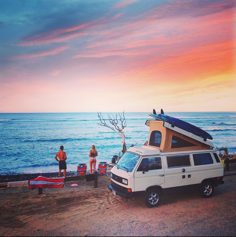 RV rental dream: White camper van with surfboards parked beachfront at stunning sunset. Couple enjoys ocean view.
