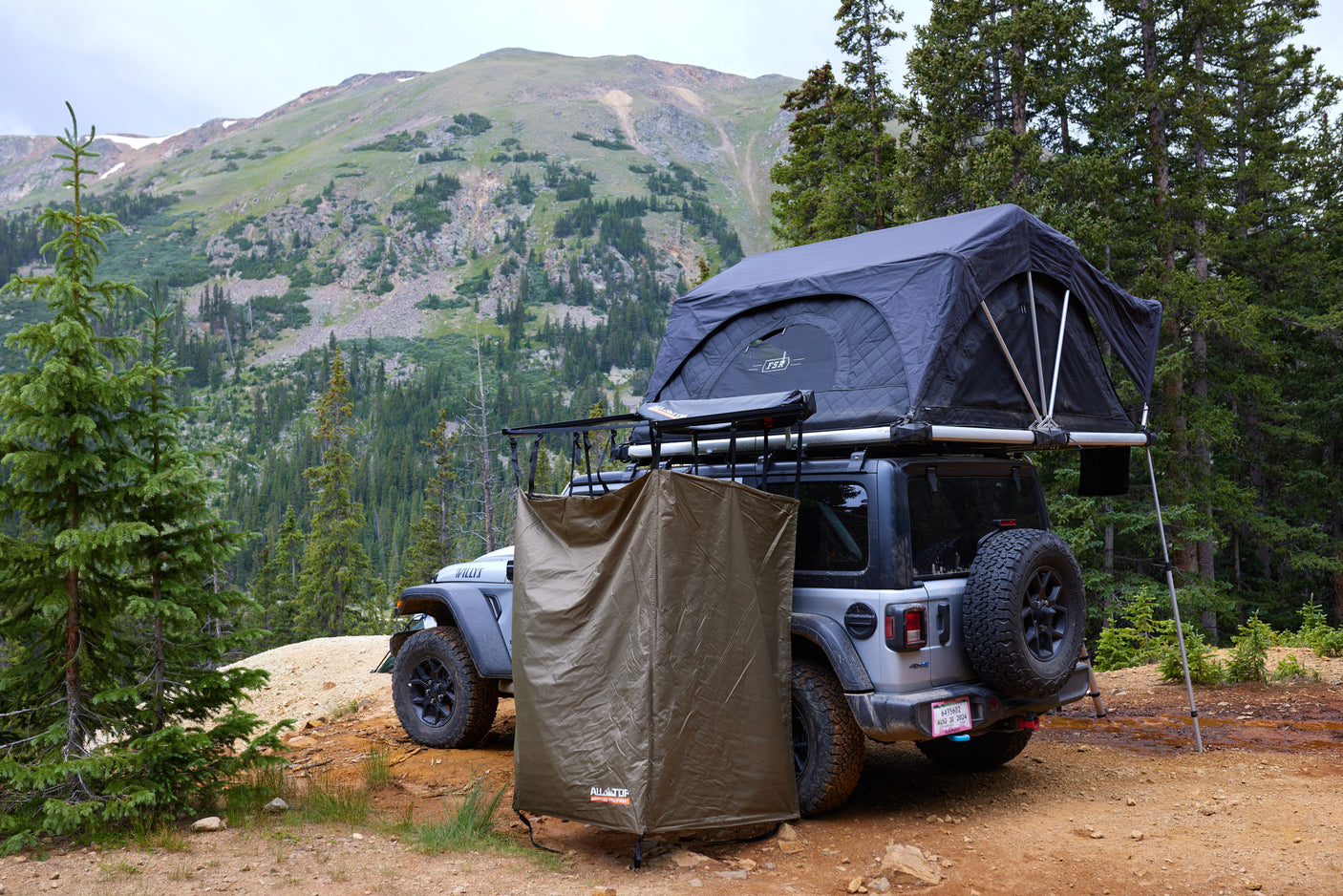 An RV rental Jeep Wrangler, equipped with a rooftop tent and privacy enclosure, parked for camping in a scenic mountain forest.