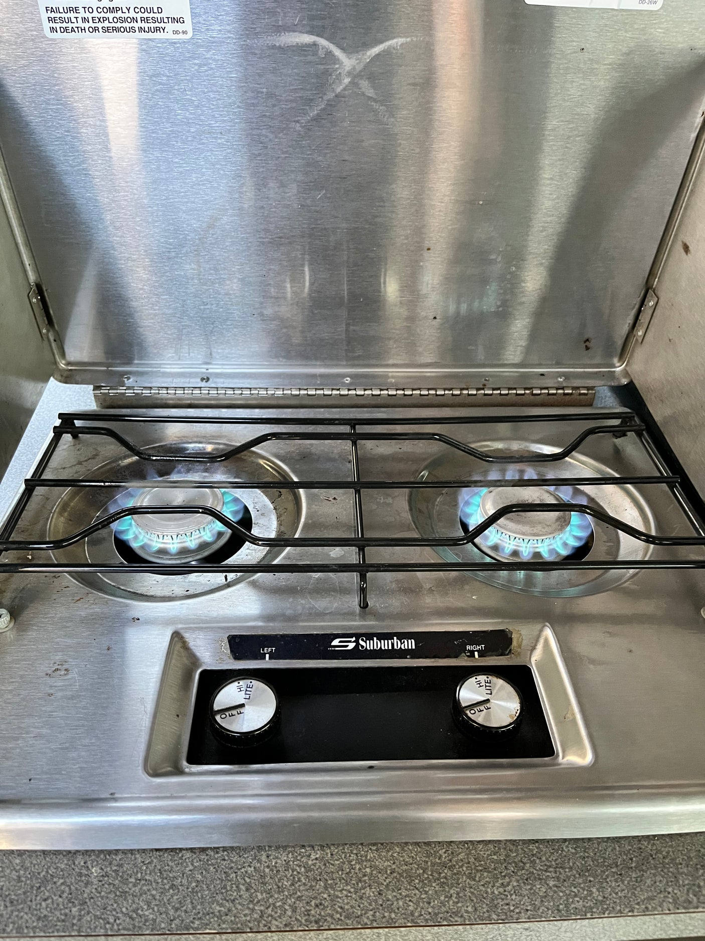An RV rental two-burner gas stove with lit blue flames, a Suburban brand, control knobs, and a visible safety warning.