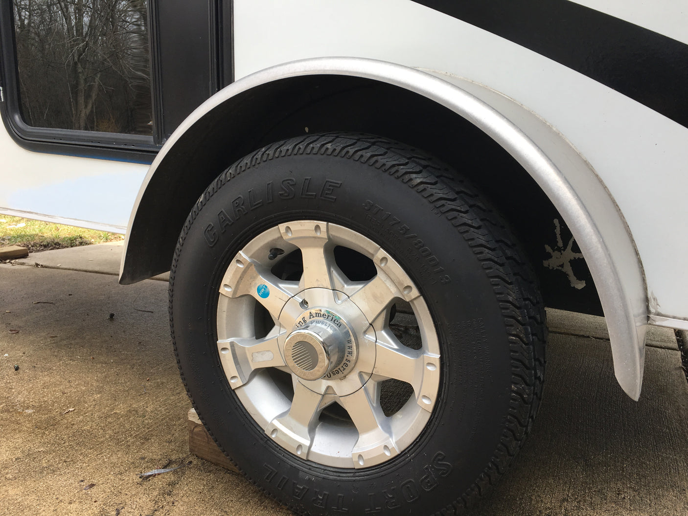 Close-up of a Carlisle ST175/80D13 tire & silver wheel on an rv rental trailer, parked with wood block.