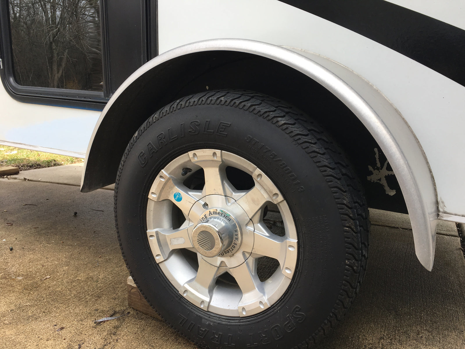 Close-up of a Carlisle ST175/80D13 tire & silver wheel on an rv rental trailer, parked with wood block.