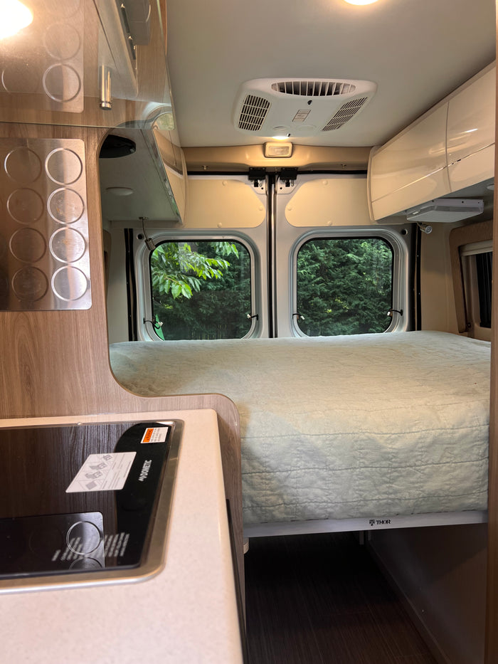 Cozy rv rental interior: view of the comfy bed, kitchen area with Dometic cooktop, and large windows with nature views.