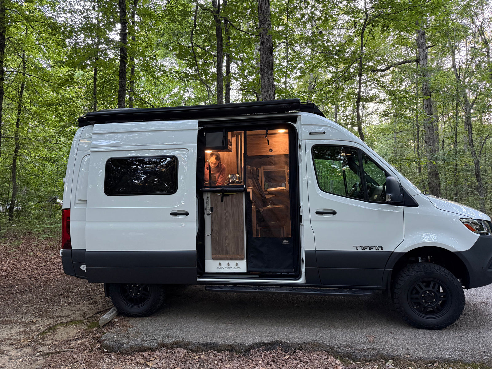 Enjoying an RV rental adventure. A Tiffin camper van with its door open, showing a woman inside working, parked in a forest.