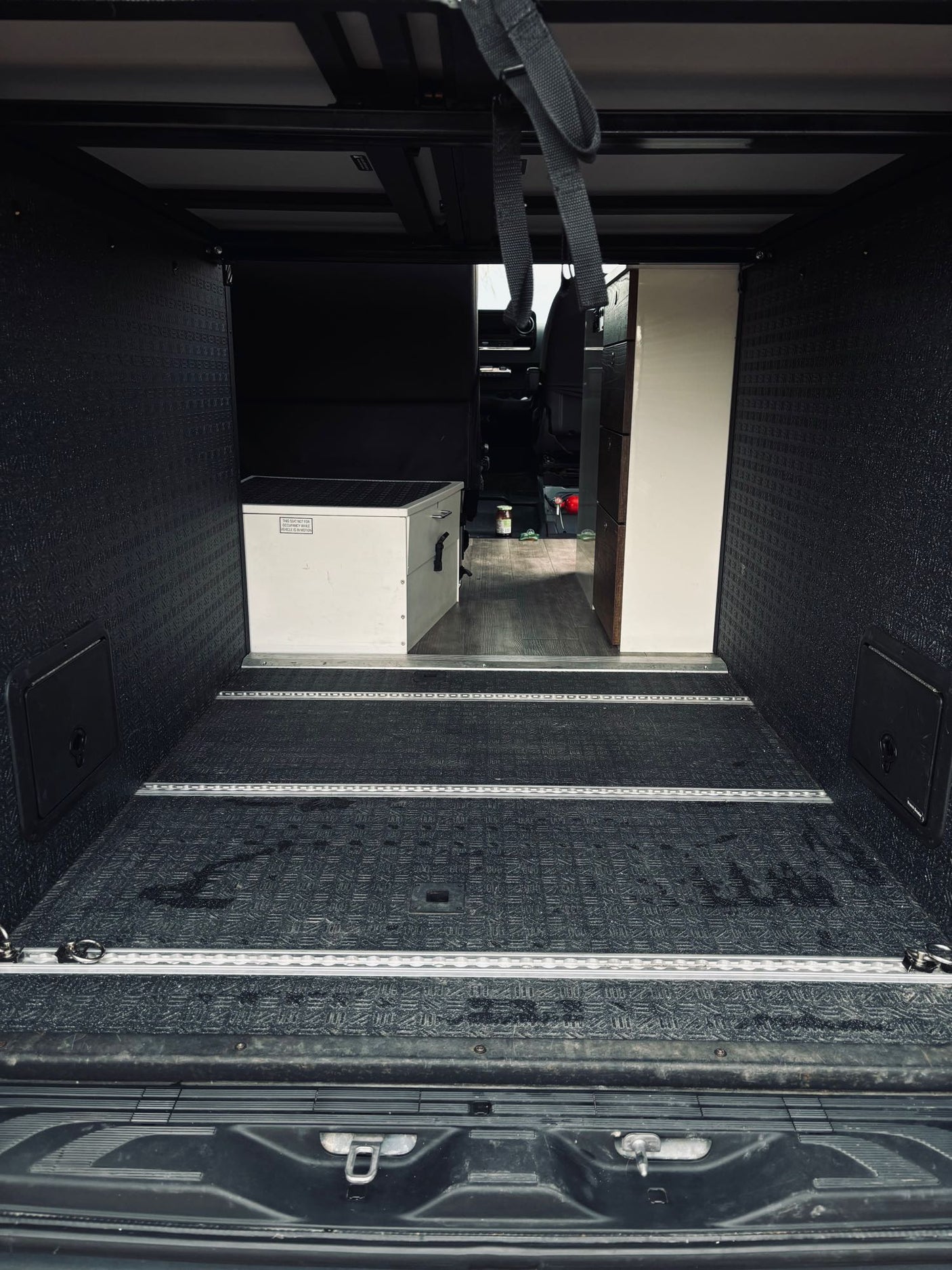 Interior of an RV rental's rear cargo space: dark, textured walls/floor, aluminum tie-down tracks, and a white storage box.