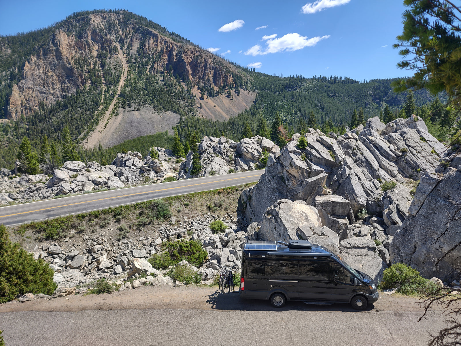 An RV rental camper van with bikes, parked on a scenic winding mountain road, amidst rocky cliffs and green forest.
