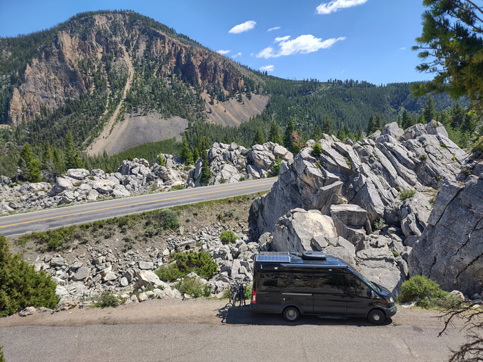An RV rental camper van with bikes, parked on a scenic winding mountain road, amidst rocky cliffs and green forest.
