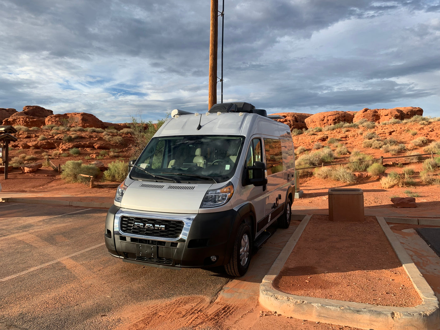 Silver RAM rv rental camper van parked in a red-dirt lot, backed by scenic red rock formations and a cloudy sky.