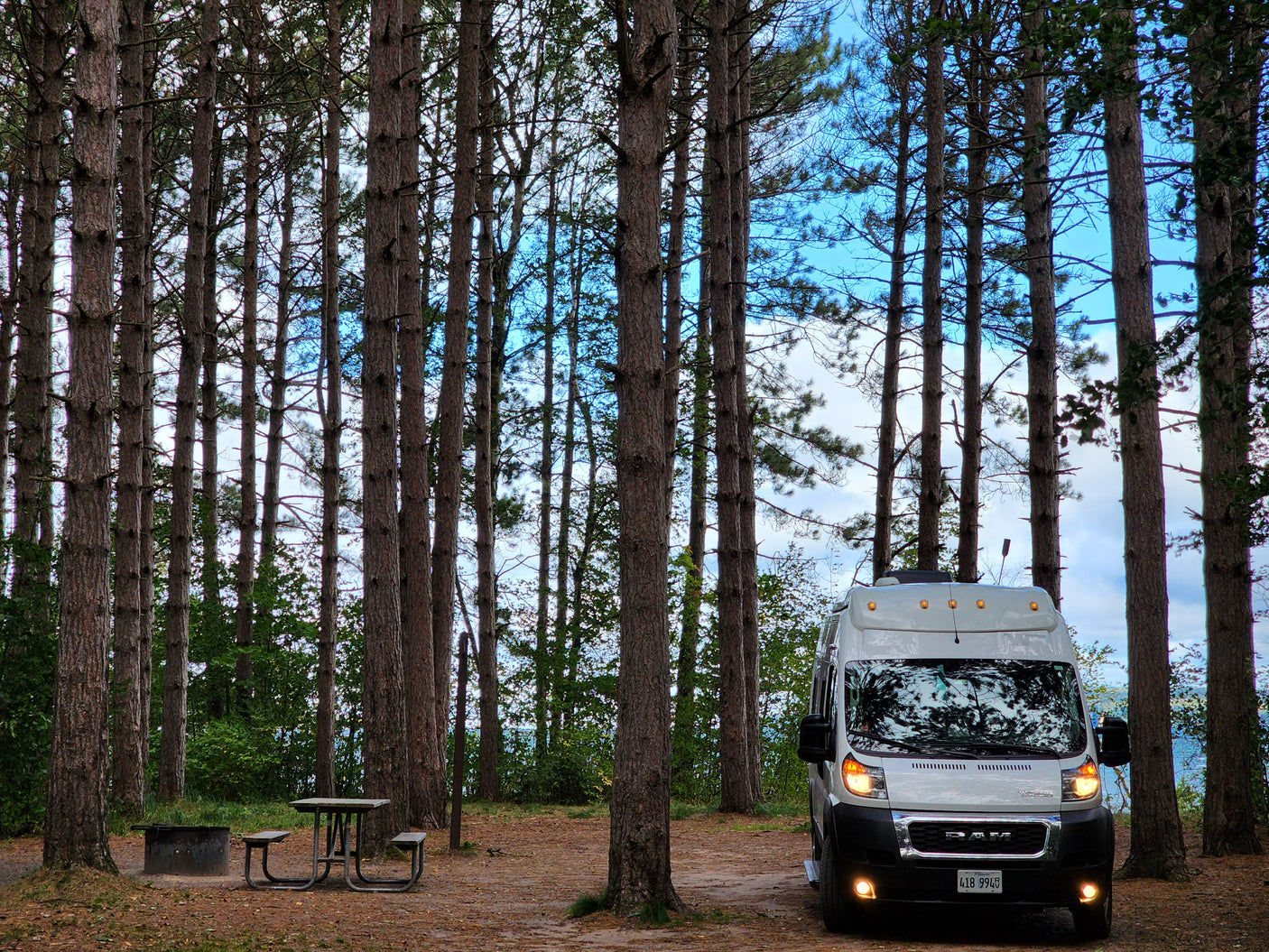 White RV rental parked among tall pine trees at a campsite with a picnic table and fire pit, overlooking distant water.