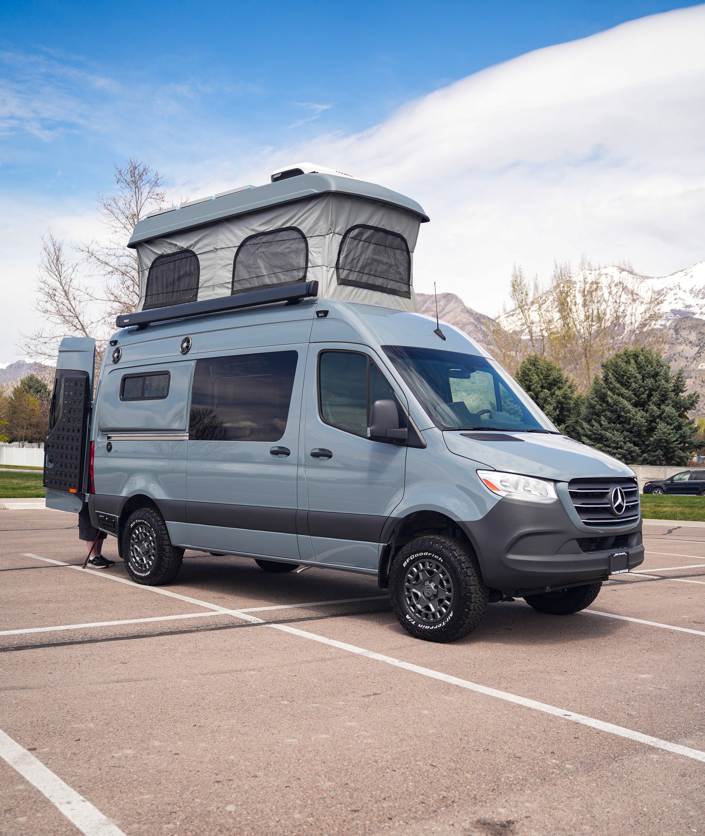 Modern gray Mercedes Sprinter pop-top camper van with off-road tires, parked in a lot. Ideal for RV rental travel.